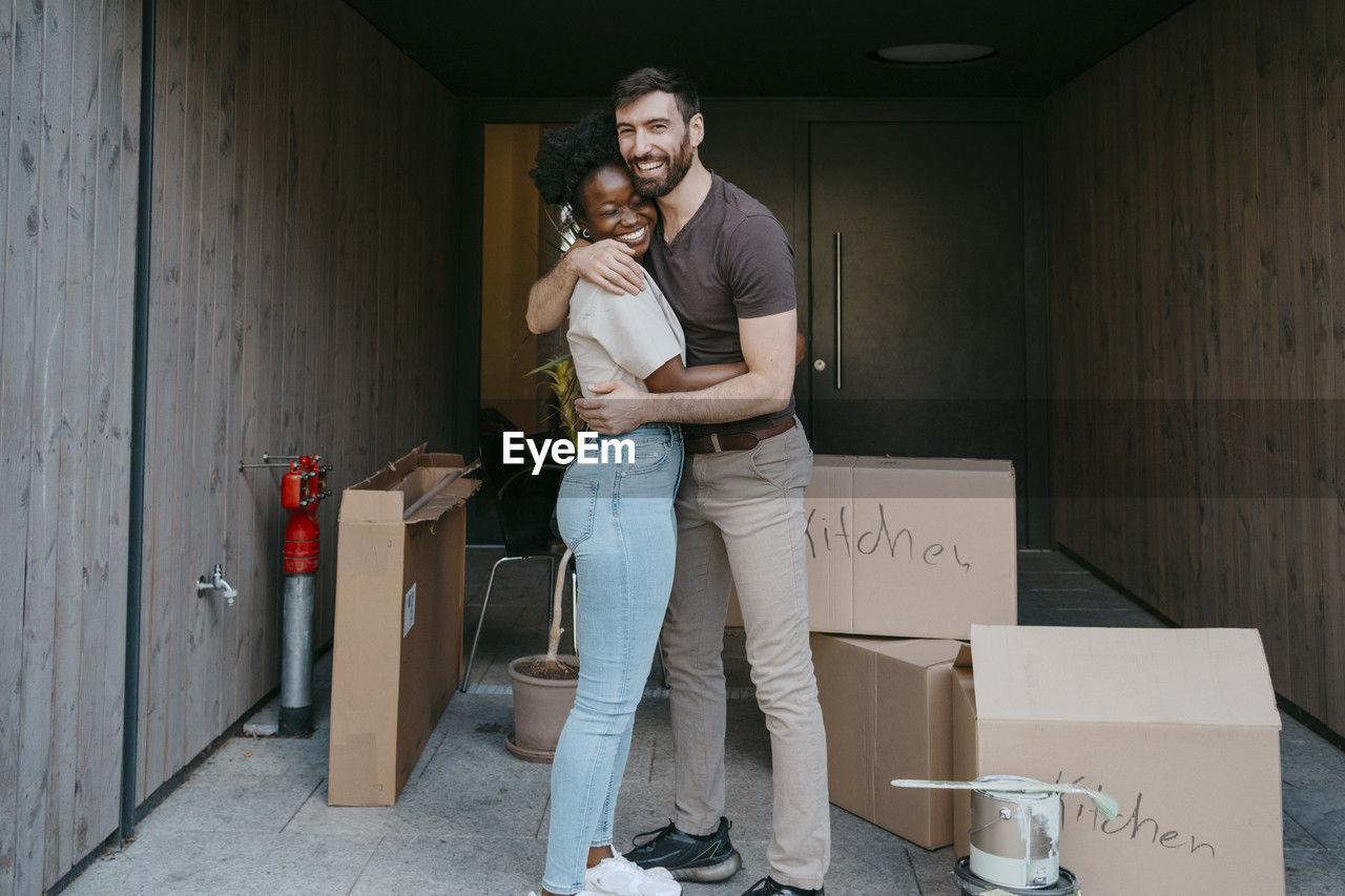 Portrait of happy man embracing girlfriend while standing amidst cardboard boxes