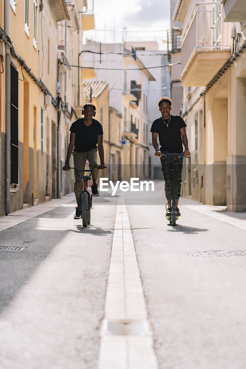 Cheerful young african american man riding electric scooter while black male is driving bicycle in street looking at camera