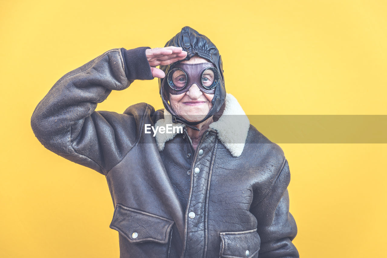 Portrait of senior woman wearing glasses while saluting against yellow background

