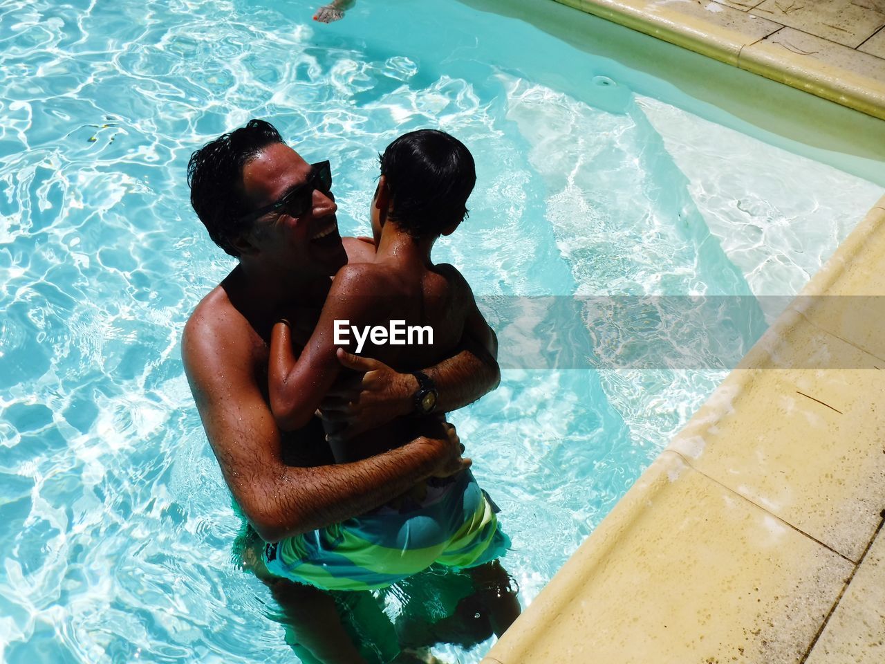 High angle view of shirtless father and son embracing in pool