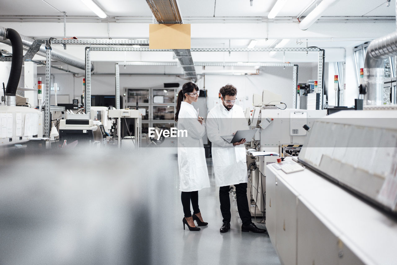 Male and female scientists discussing over laptop by machinery at laboratory