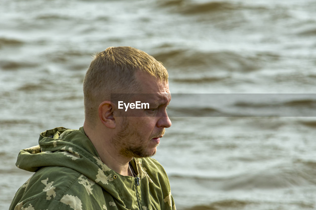 Unshaven fisherman against background of waves on river looks forward. side view.