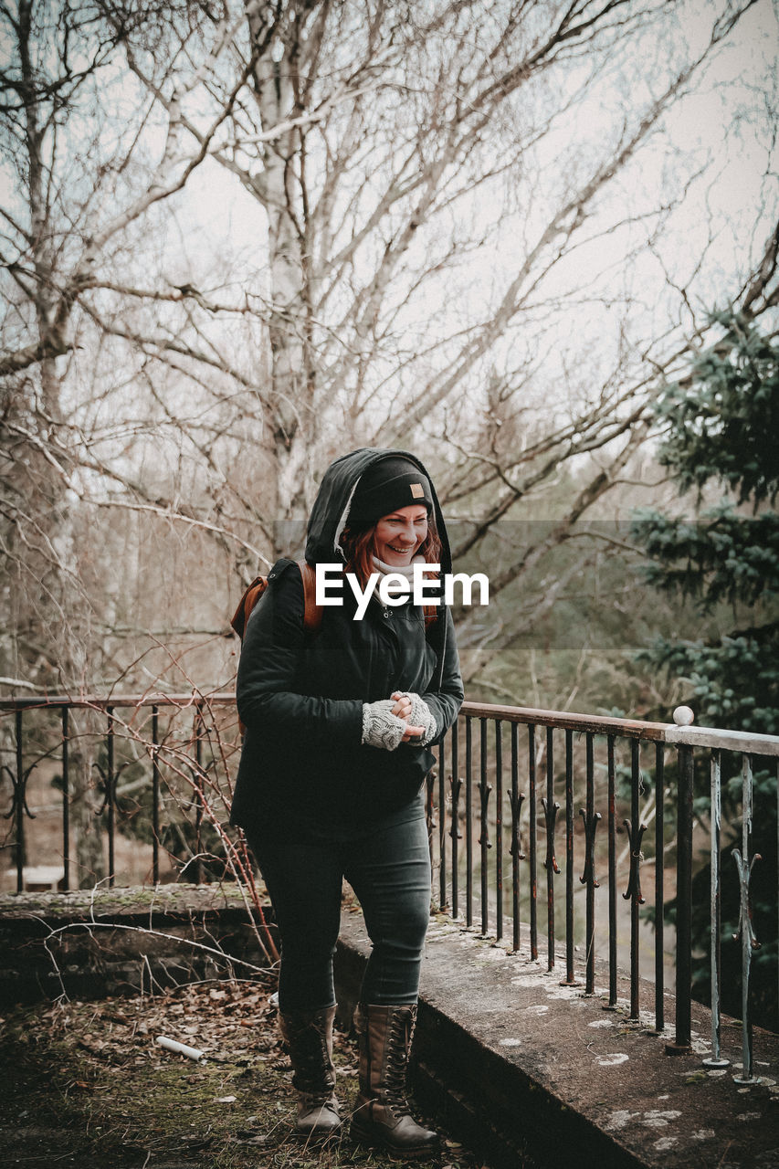 one person, tree, spring, full length, clothing, lifestyles, winter, adult, nature, women, young adult, standing, railing, leisure activity, bare tree, plant, cold temperature, warm clothing, front view, day, casual clothing, black, portrait, outdoors, fence, footwear, emotion, architecture, female, forest, looking at camera
