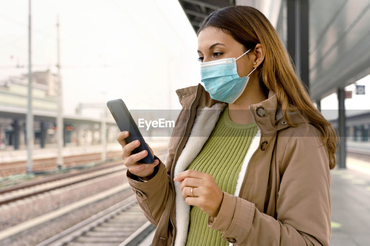 Woman with surgical mask using her smart phone waiting her train at station outside