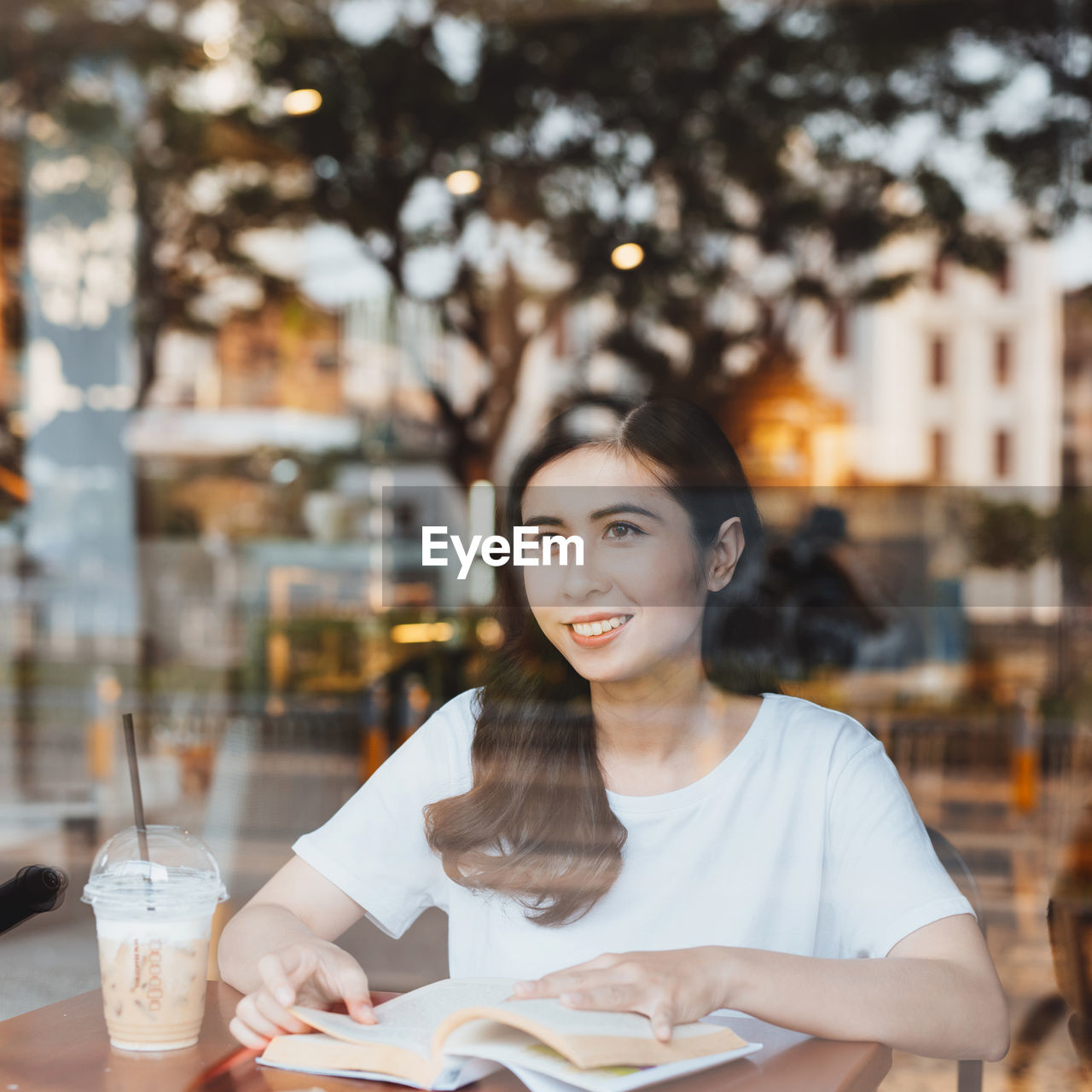 Smiling woman with coffee cup and book on table seen through glass window