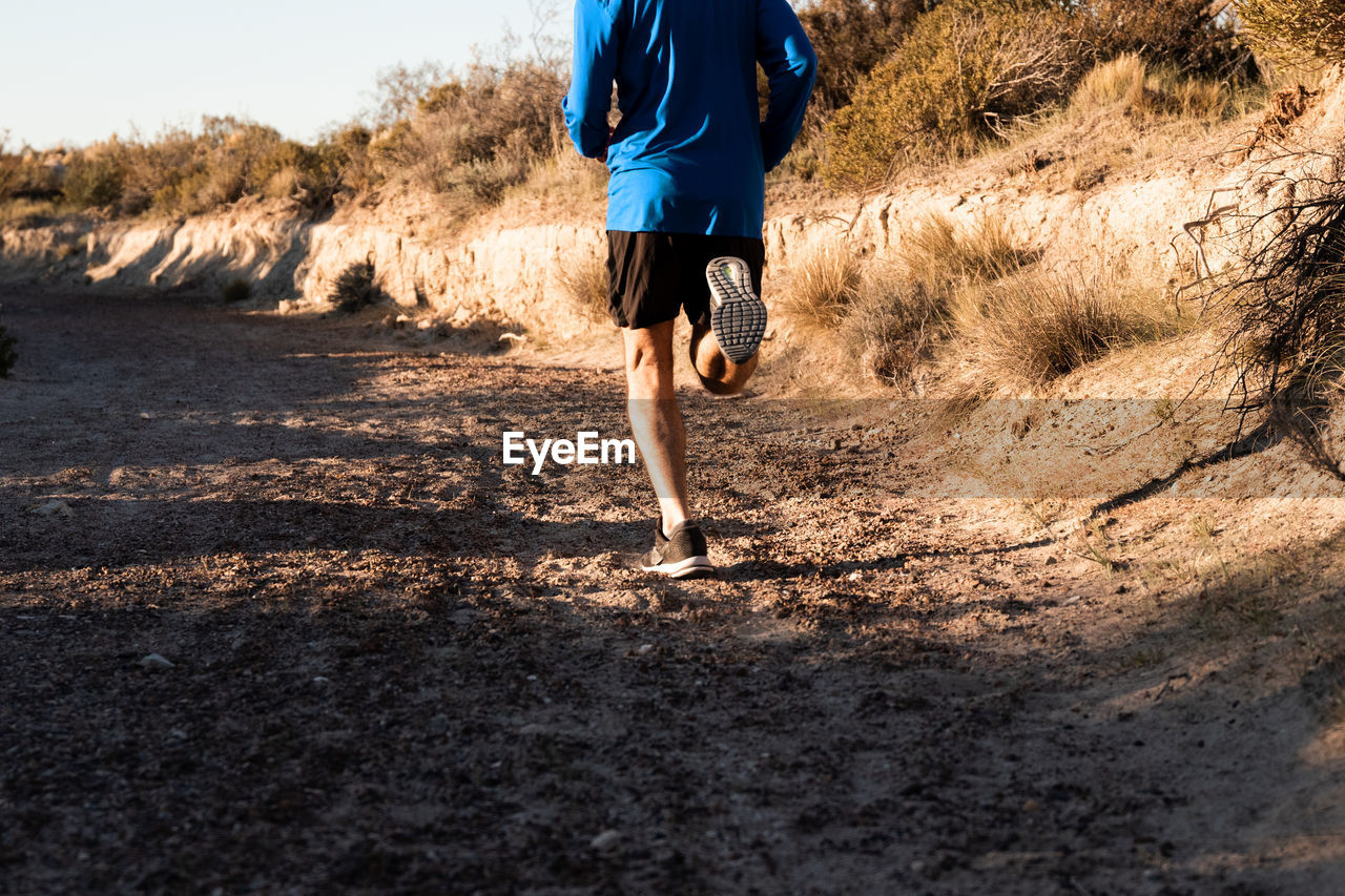 Back view of unrecognizable active male jogger running uphill in sandy semi desert terrain during workout in sunny morning