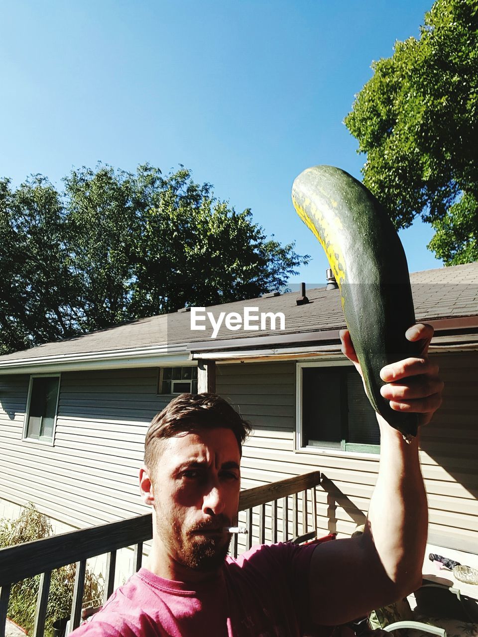 Portrait of man holding cucumber while smoking cigarette in front of house
