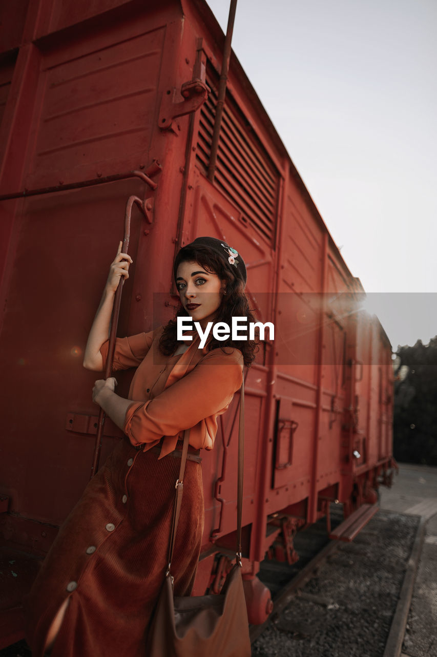 Young female with dark curly hair in beret wearing terracotta clothes in vintage style in back lit standing on step of car train painted in terracotta color and holding handrail while looking at camera flirty