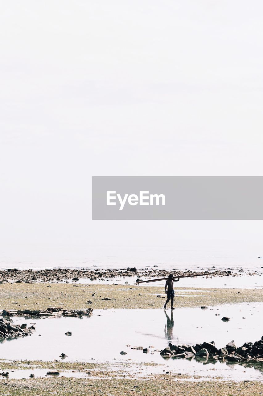 Mid distance view of shirtless man carrying wood on shoulder at beach against clear sky