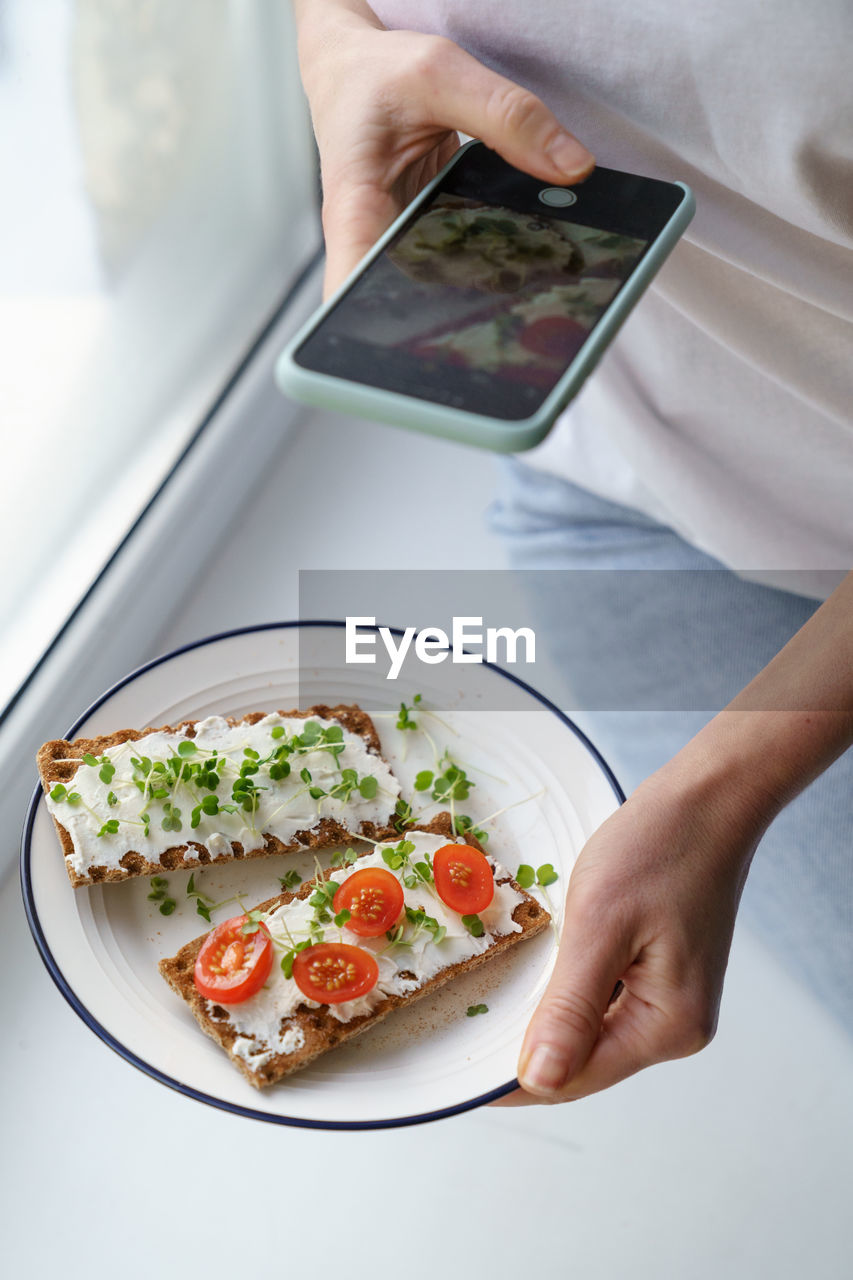 Woman takes a photo on cellphone hold plate with rye crisp bread with creamy cheese and micro greens