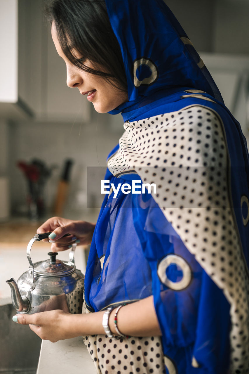 Young woman holding teapot while standing in kitchen at home