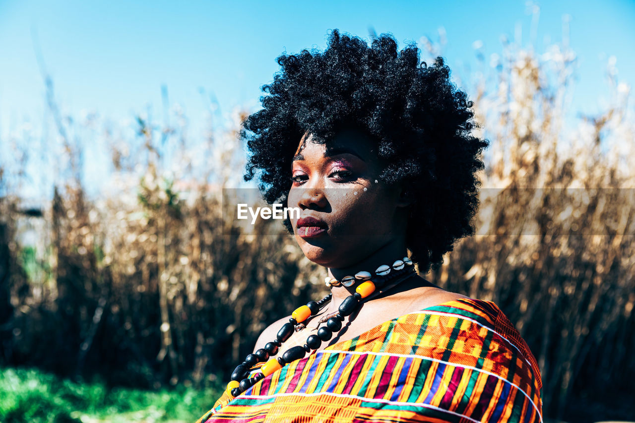 Serious african model with afro hairstyle wearing colorful handwoven kente and looking at camera while standing on field on bright summer day