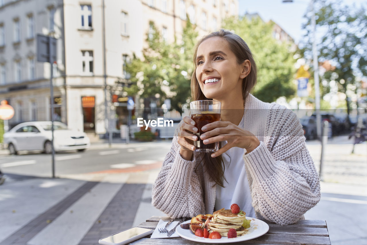 Smiling young female with pancake drinking while sitting at table in city