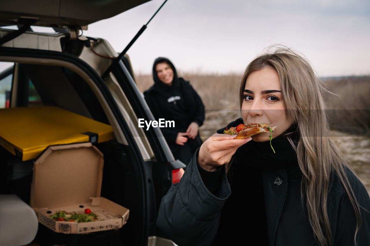 Portrait of young woman eating pizza and standing by car on the beach