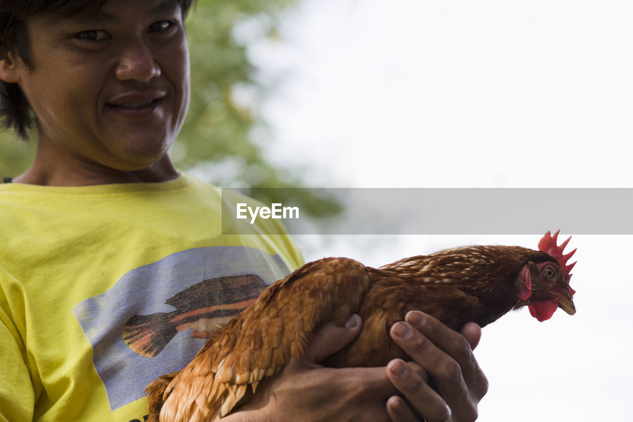Low angle portrait of farmer holding hen while standing against clear sky