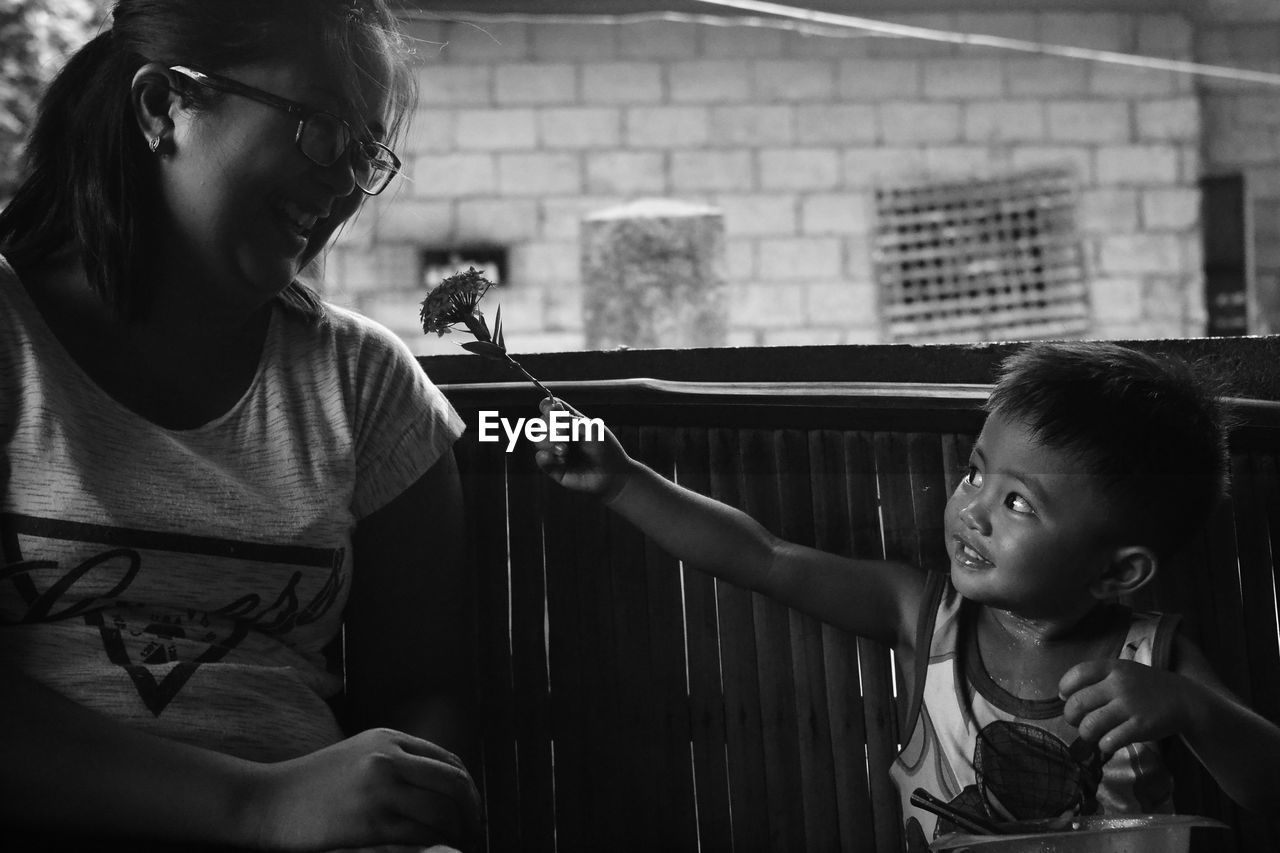 Boy giving flower to mother while sitting on sofa at home