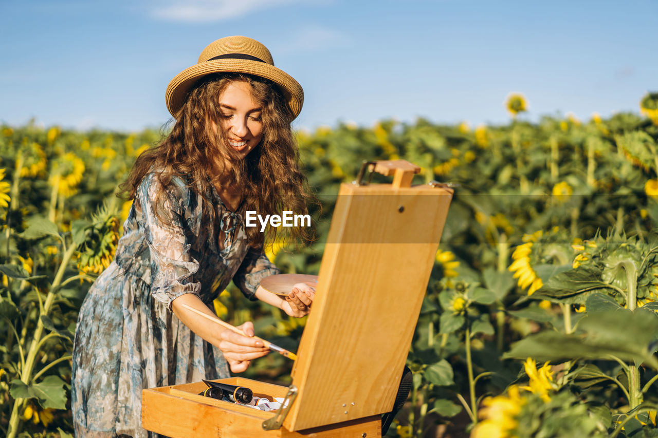 A young woman with curly hair and wearing a hat is painting in nature. 