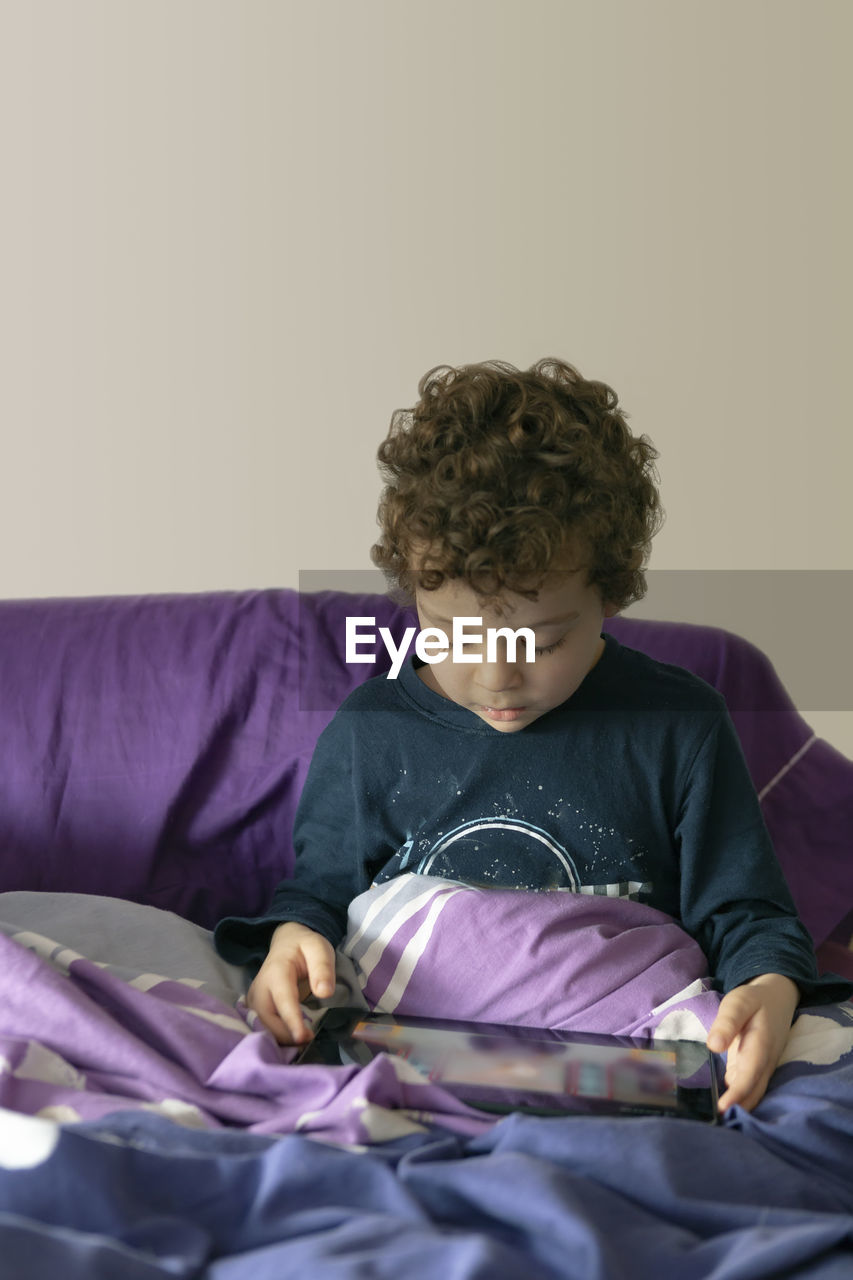 4-5 year old boy with curly hair sitting on the bed with a tablet in his hands playing 