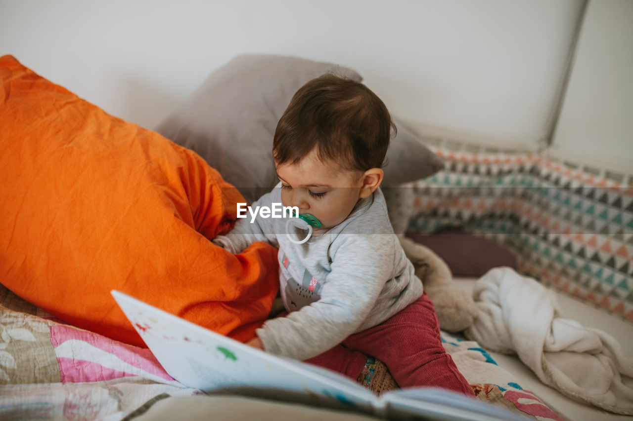 Cute baby boy reading book while sitting on bed at home