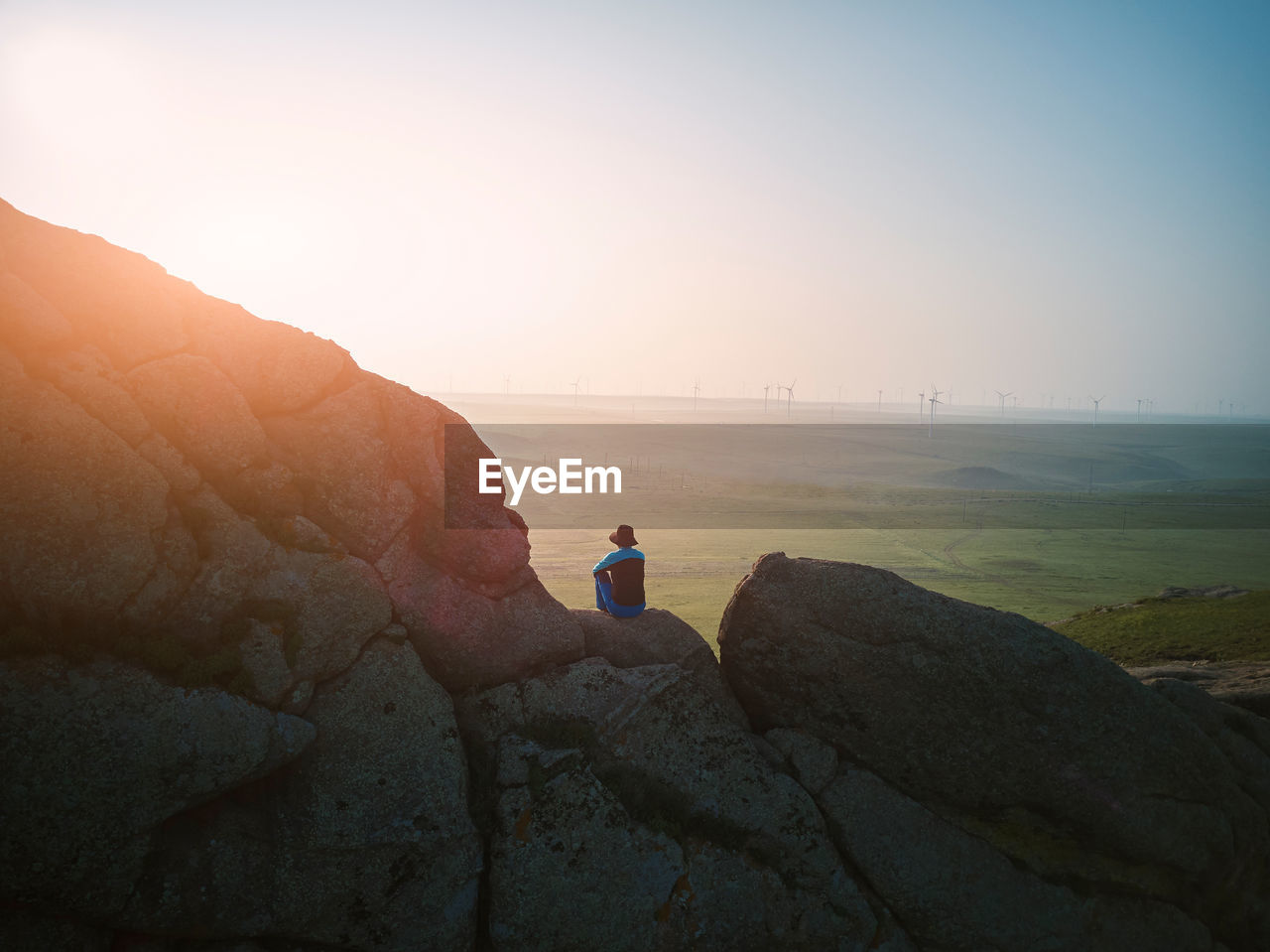 Rear view of man sitting on cliff against sky during sunset