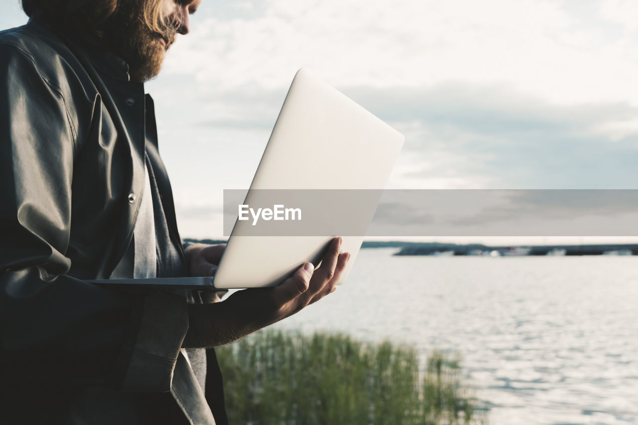 Midsection of man using laptop by lake against sky