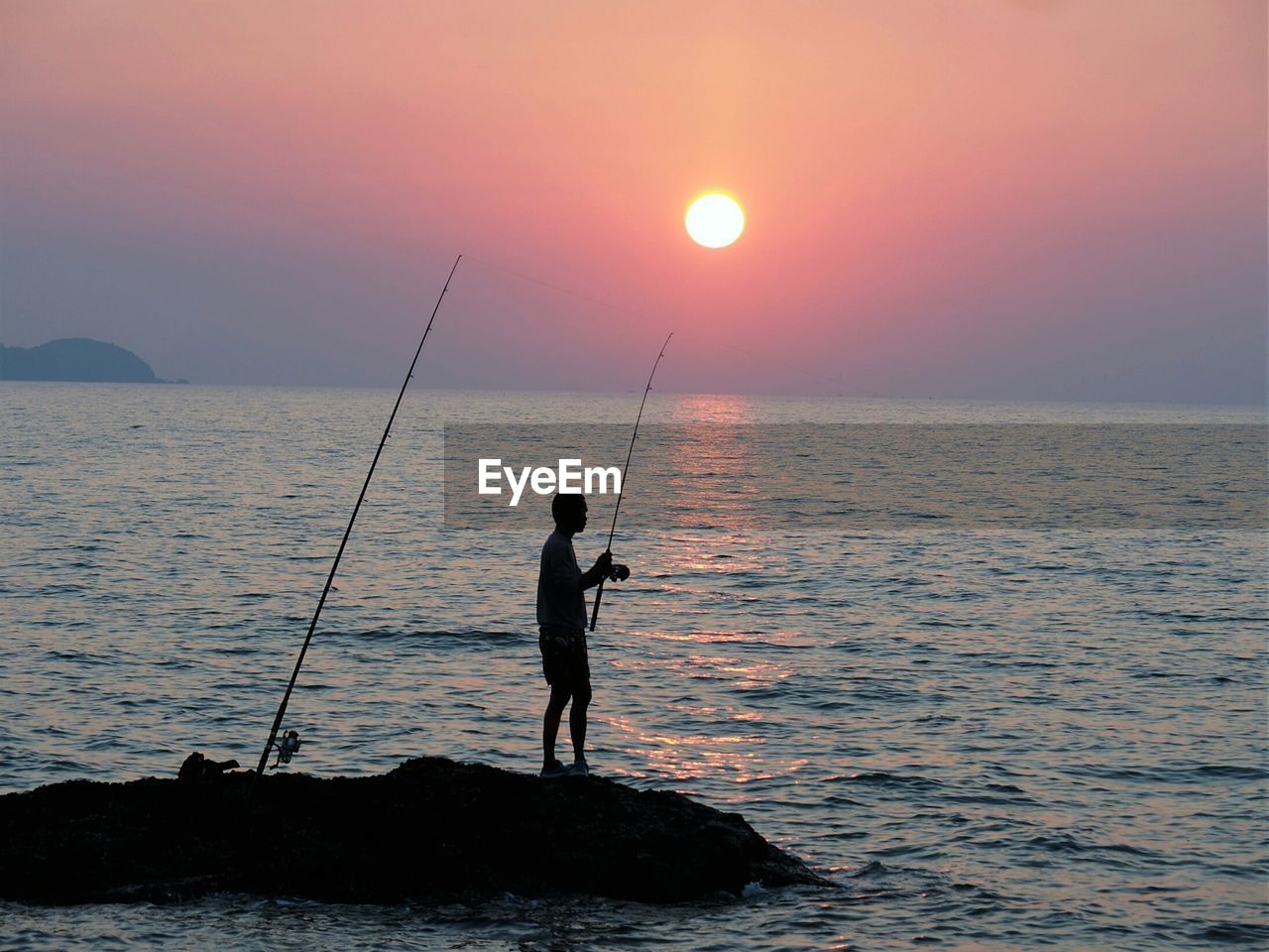 Man standing on rock formation while fishing in sea against sky during sunset