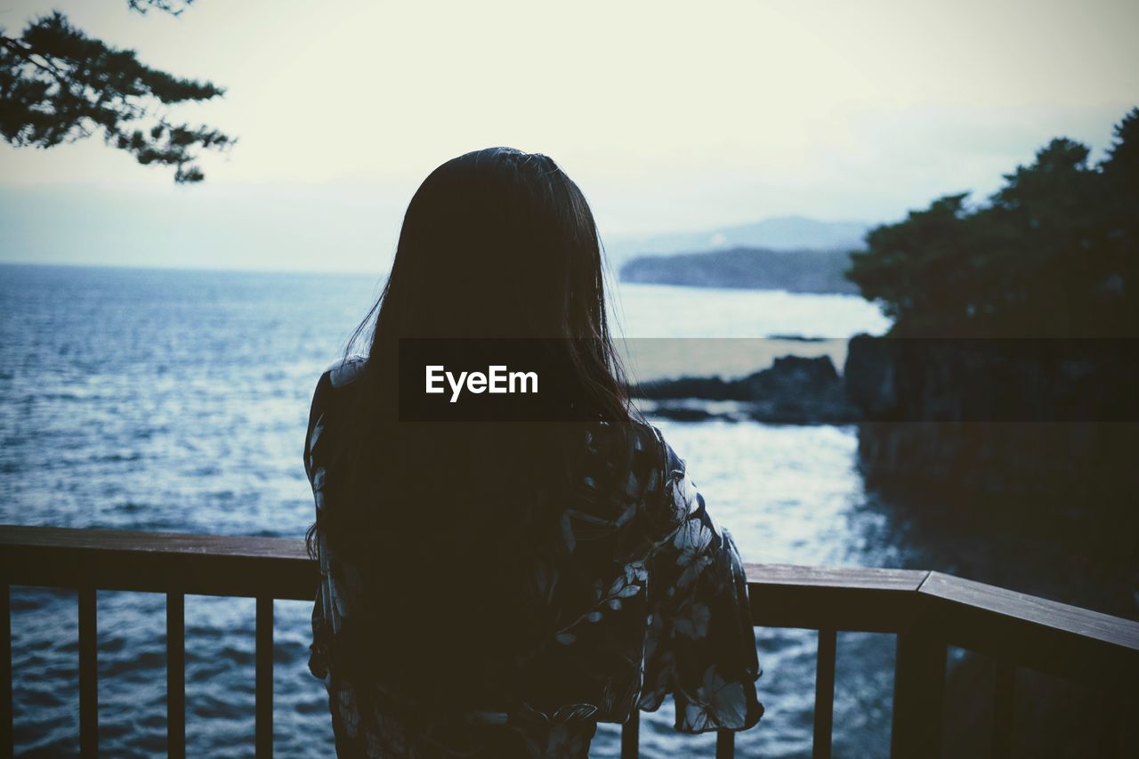 REAR VIEW OF WOMAN LOOKING AT SEA SHORE AGAINST SKY