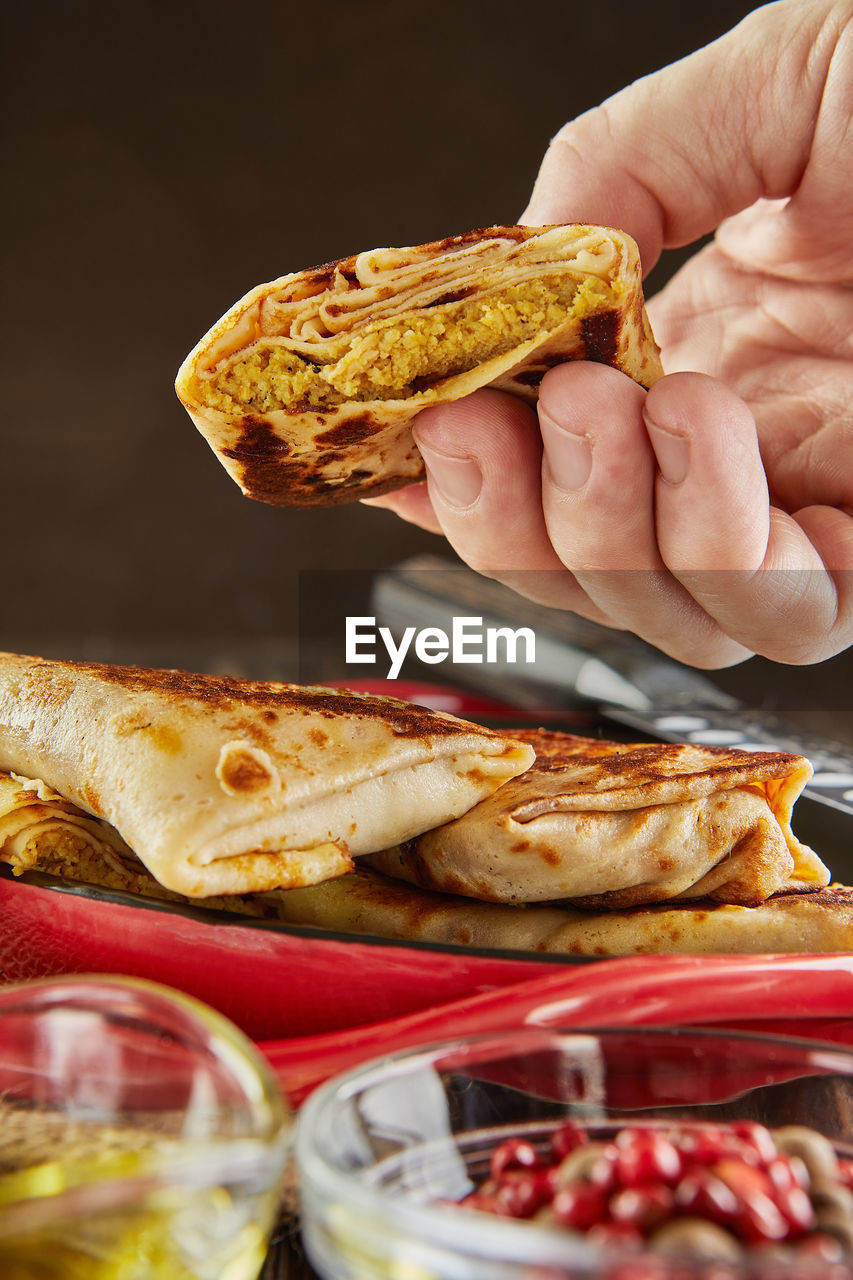 Pancakes with meat, holds by hand, with oil and tongs on plate on dark wooden background