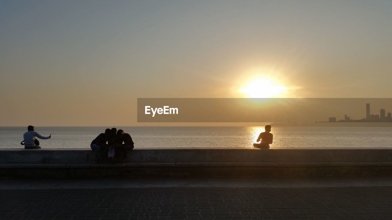People sitting on retaining wall by sea against sky during sunset