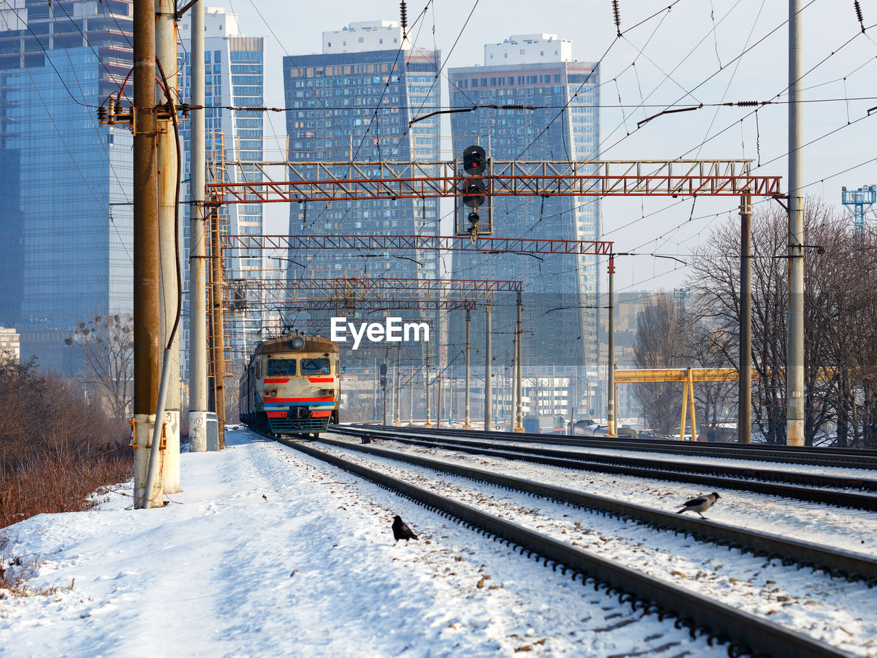 An old electric train moves on rails in the winter season against the backdrop of a cityscape.