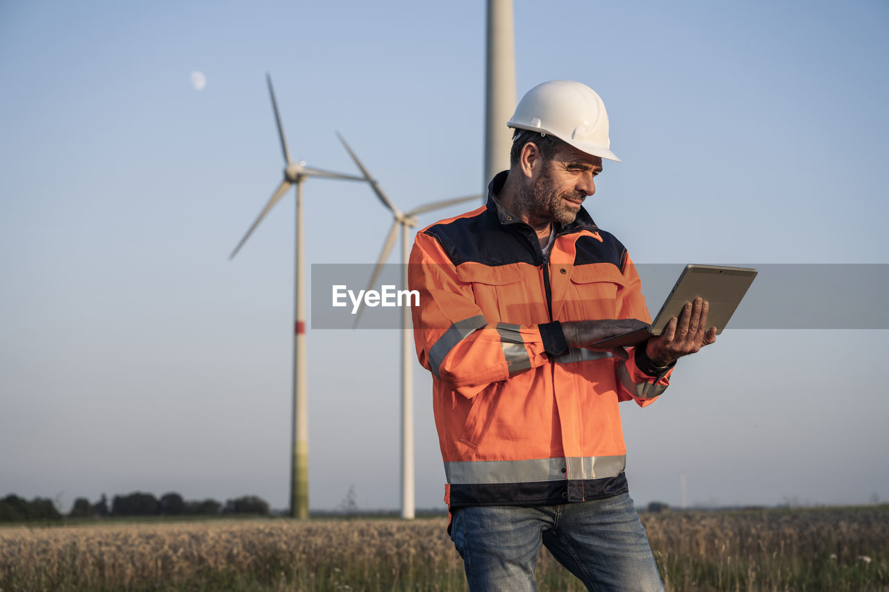 Male engineer using digital tablet while standing in front of wind turbines at sunset