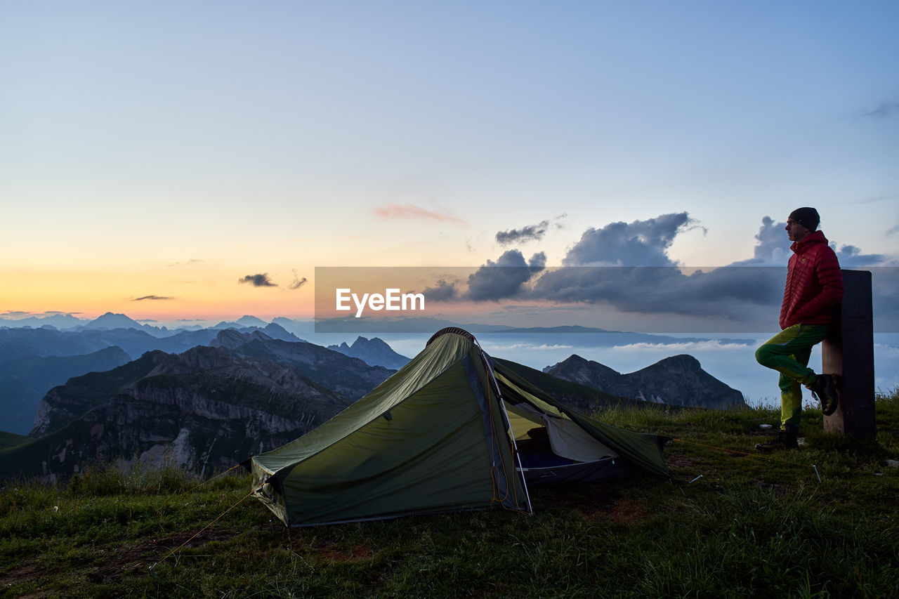 Full length of man looking at view while standing on mountain by tent against sky during sunset