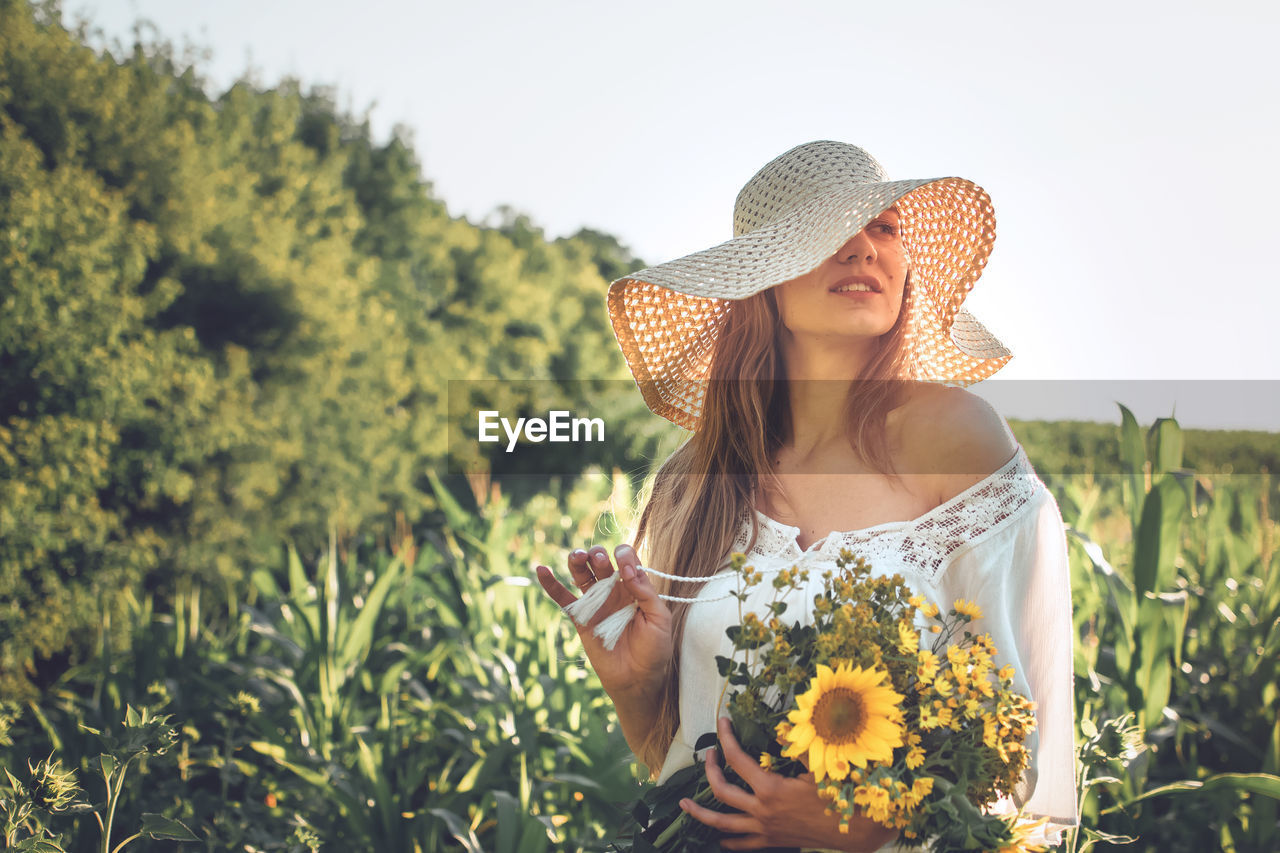 Woman holding flower while standing against sky