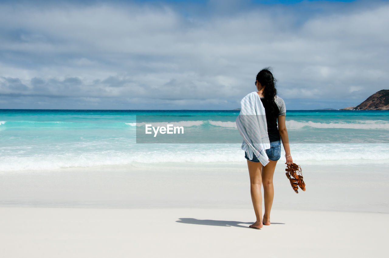 Rear view of mid adult woman standing at beach against cloudy sky