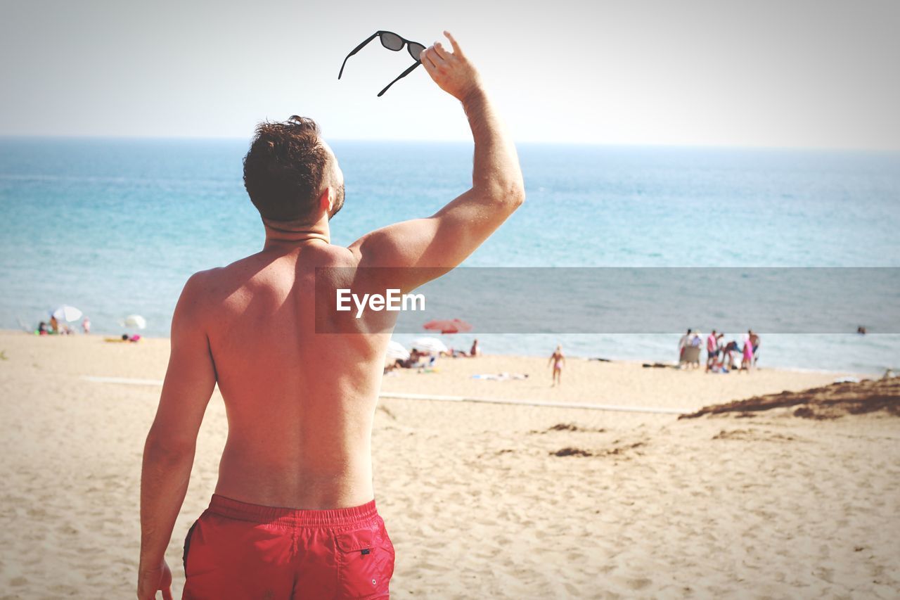 Rear view of shirtless mid adult man holding sunglasses while standing at beach against clear sky