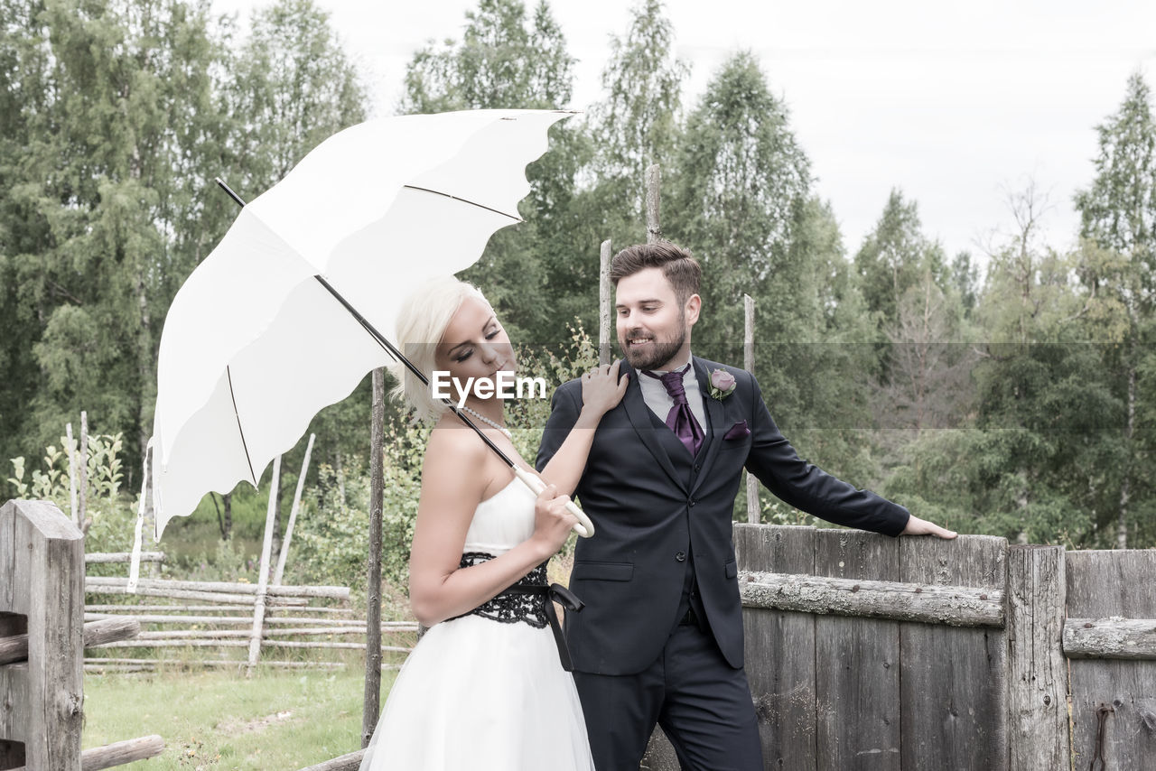 Bride with groom holding umbrella against trees