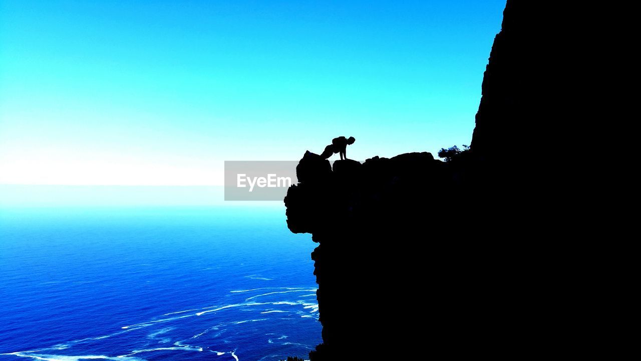 Silhouette man exercising on cliff by sea against clear blue sky