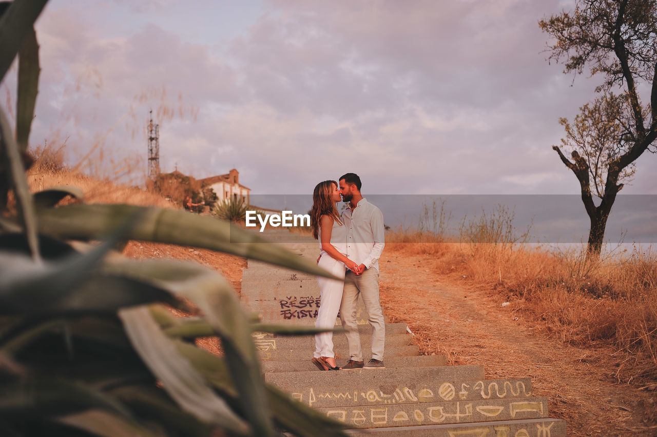 Full length of young couple kissing on steps against cloudy sky