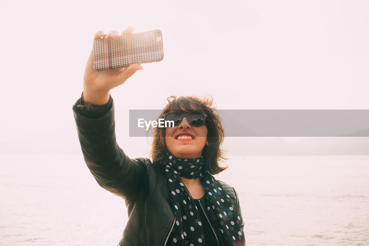 Smiling woman taking selfie by sea against clear sky