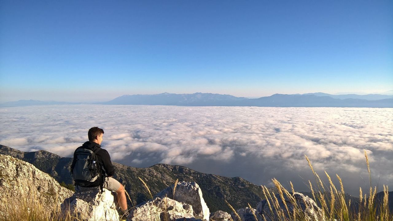 Rear view of man sitting on mountain against cloudscape