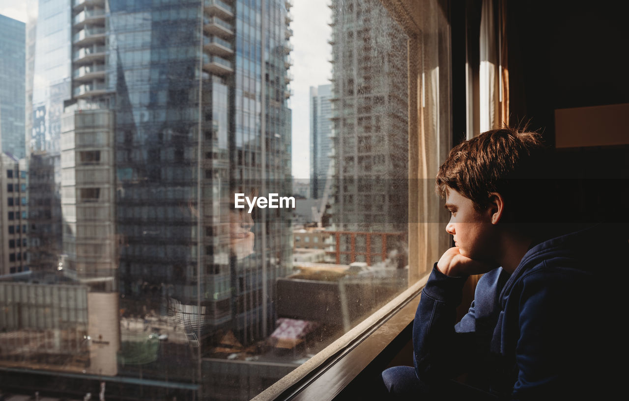 Tween boy looking out a window at tall buildings of the city outside.