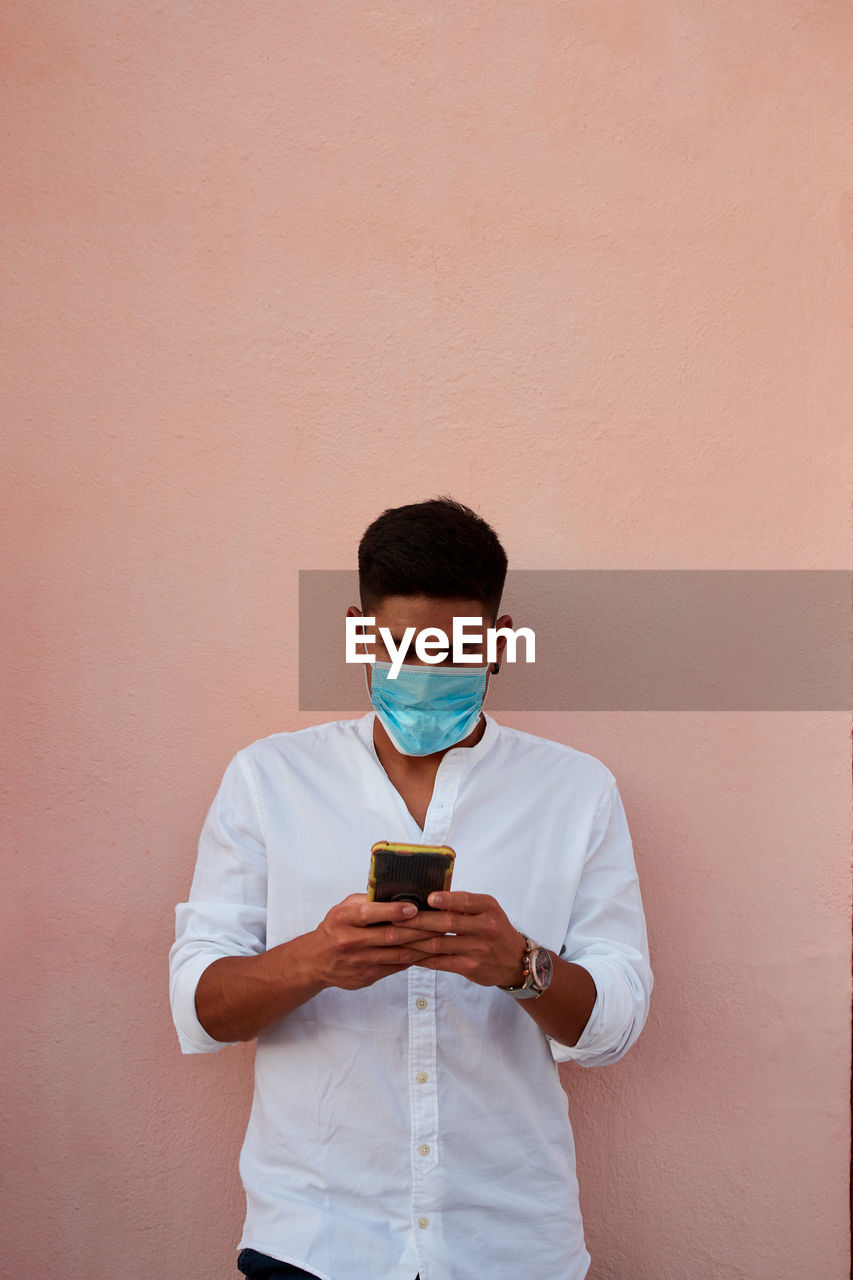 Young latin man with mask is looking his phone on pink background