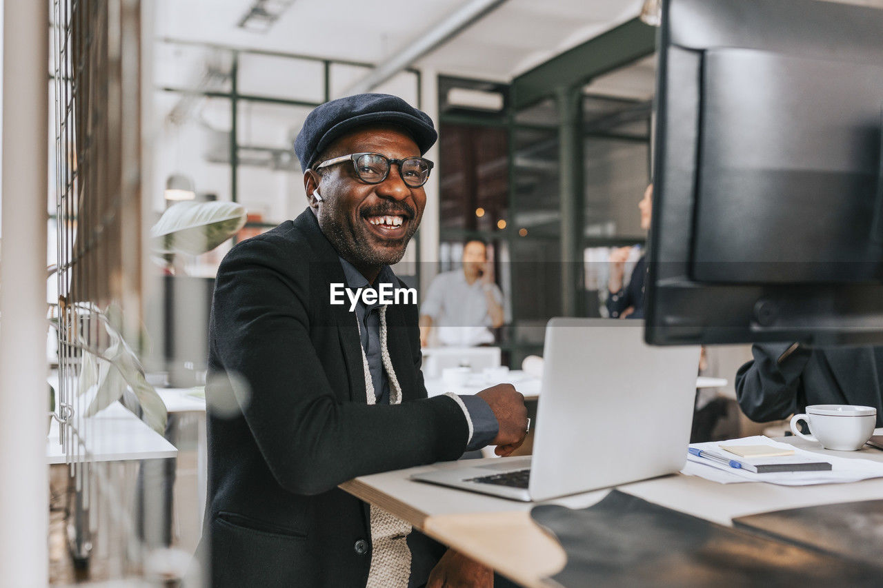 Happy mature businessman wearing wireless in-ear headphones sitting with laptop at office