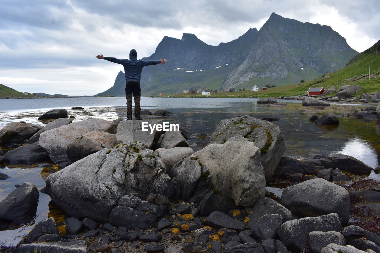 REAR VIEW OF MAN STANDING ON ROCKS AT SHORE AGAINST SKY
