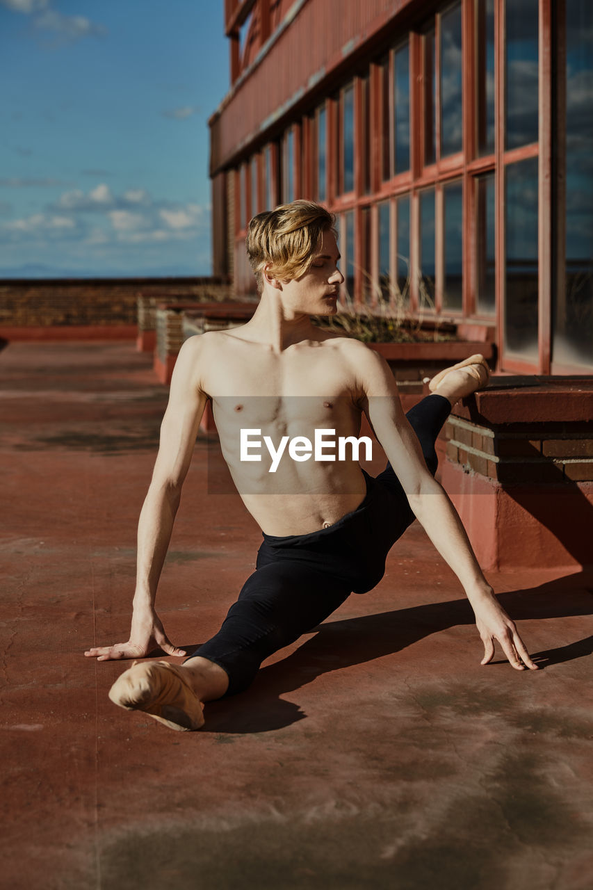 Concentrated young blond slender ballet dancer in black leotard and pointe shoes performing transverse twine on roof of building in sunny day and looking away