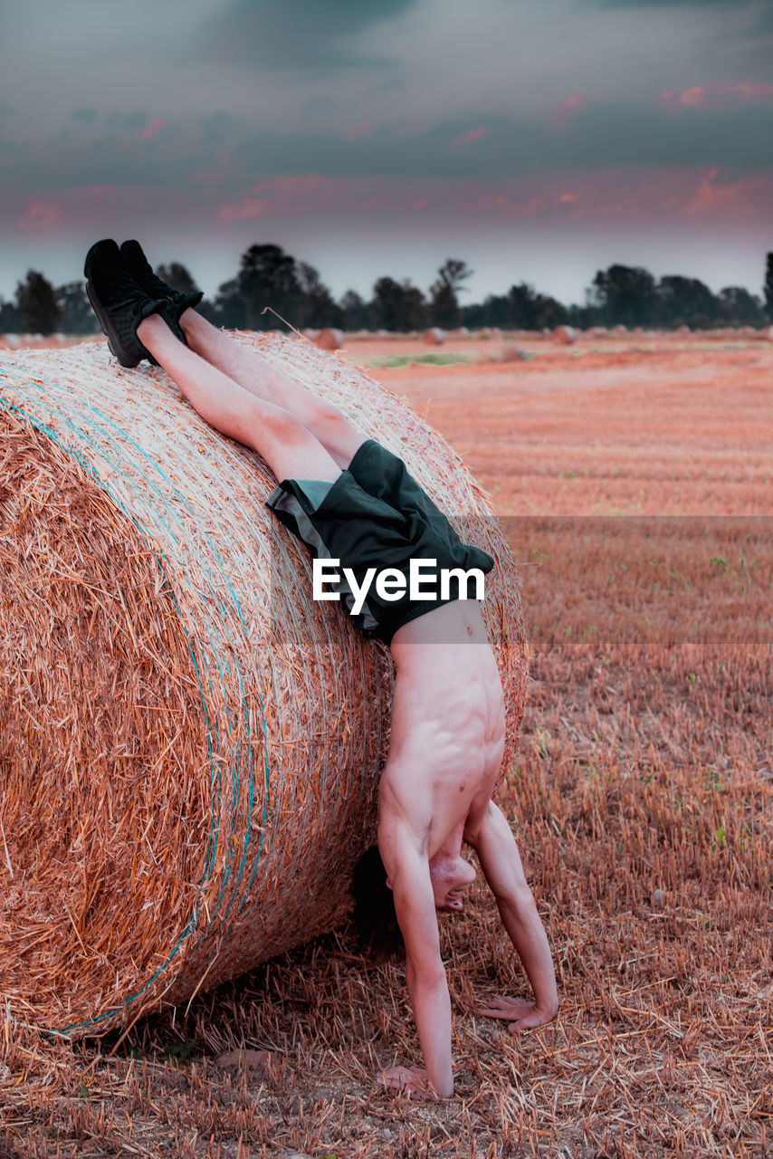 Shirtless teenage boy doing handstand by hay bale at farm
