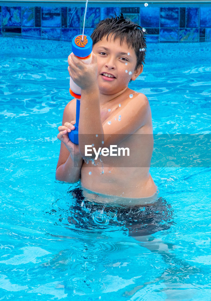 A young boy with a colorful water gun, sprays his brother as he splashes in a pool