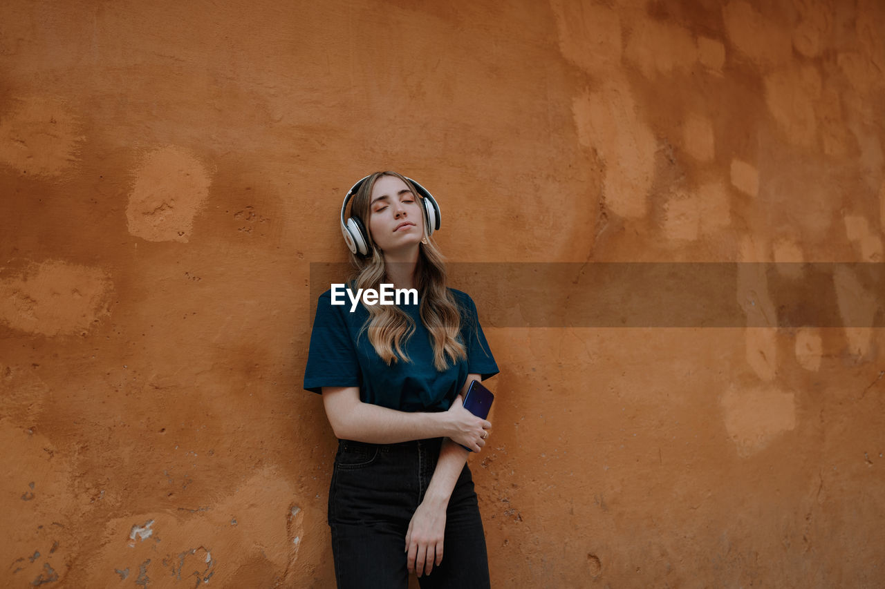 Young mindful blonde female with closed eyes listening to song in wireless headset on brown background in city