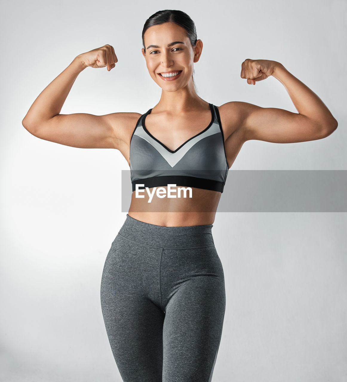 arm, women, one person, adult, exercising, portrait, lifestyles, studio shot, wellbeing, indoors, young adult, muscular build, limb, sports clothing, trunk, looking at camera, sports, vitality, undergarment, athlete, standing, smiling, strength, flexing muscles, clothing, human leg, female, sports training, happiness, gray, body conscious, physical fitness, bodybuilding, emotion, human muscle, human limb, person, front view, relaxation, gray background, cheerful, body care, slim, photo shoot, brown hair, arms raised, weight, brassiere, fitness professional, positive emotion, cut out, trousers, white background, sports bra, tank top, ponytail, enjoyment, hand