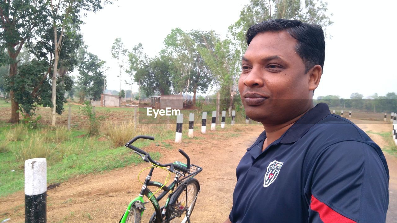 PORTRAIT OF YOUNG MAN WITH BICYCLE STANDING AGAINST TREES